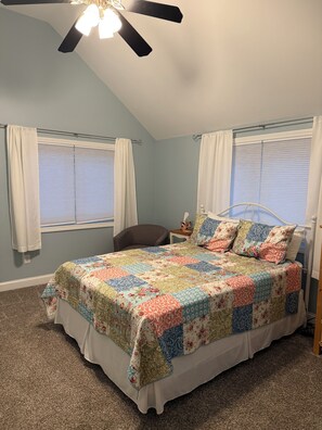 A bedroom with a bed, a chair, a nightstand, a ceiling fan, and two windows with curtains.