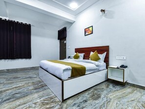 A hotel room with a bed, a bedside table, a nightstand, a wall-mounted light, and a window with curtains. - Hotel Shiv Kalpana (Jaipur)