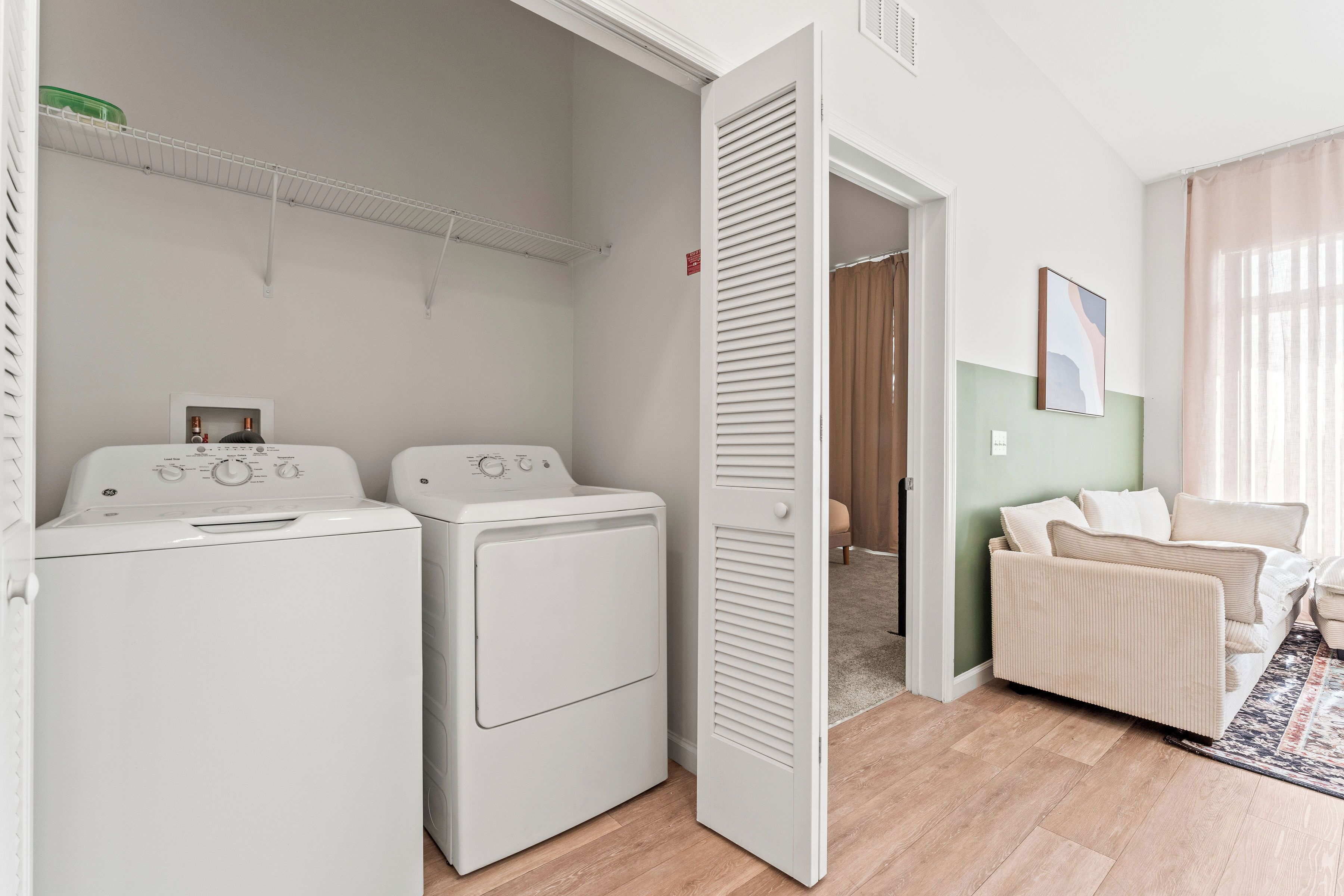 A laundry room with two white dryers, a white door, and a beige sofa in a living area with a rug.