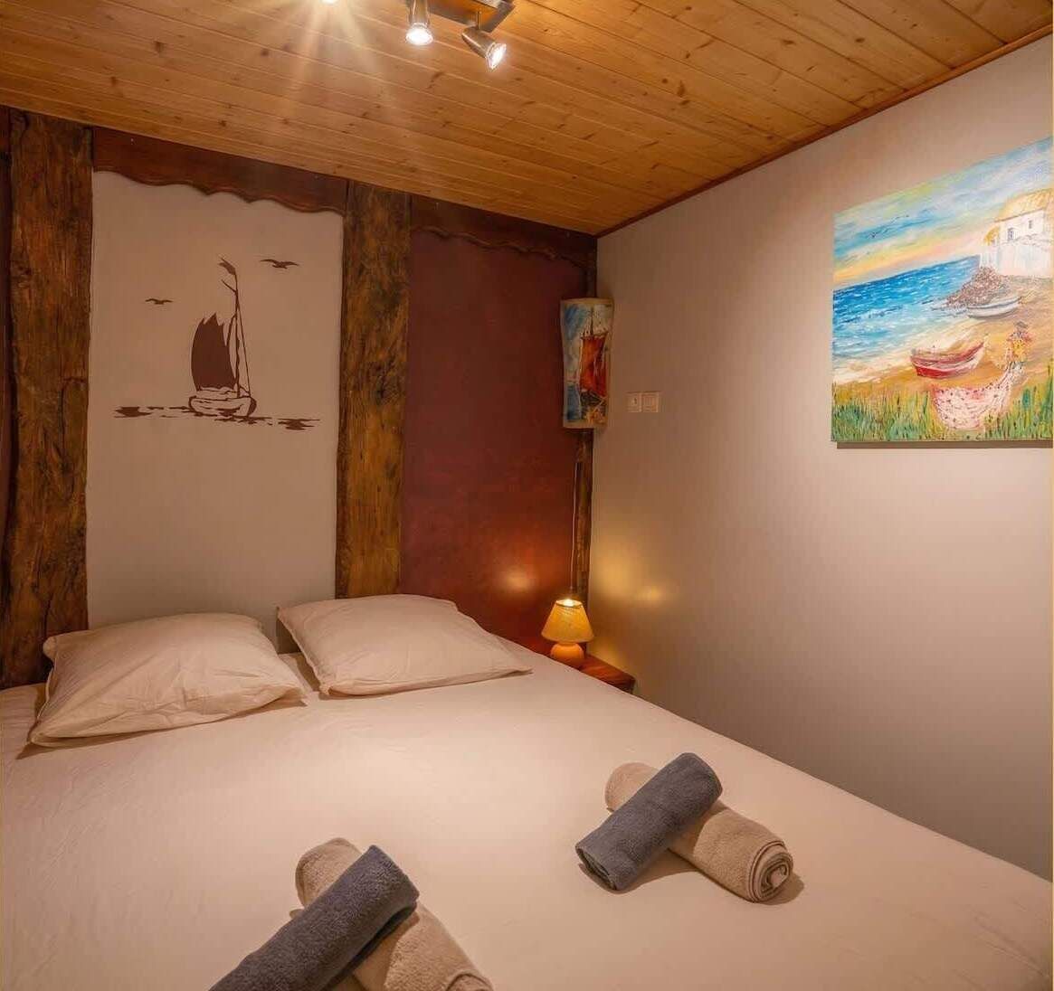 Ein Schlafzimmer mit Holzbalkendecke, einem Bett mit zwei Kissen, einem Nachttisch mit Lampe und einem Gemälde an der Wand.