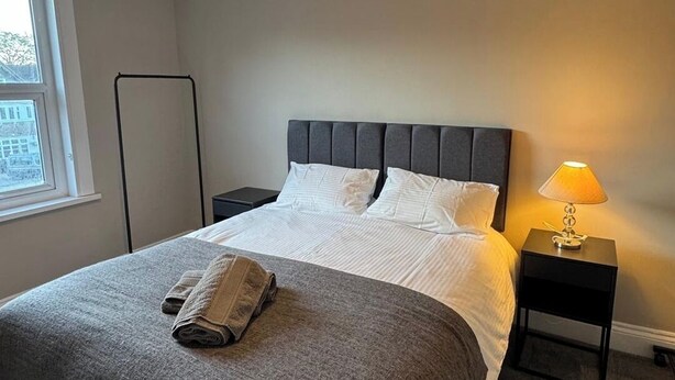 Una cama bien hecha con cabecera gris y ropa de cama blanca, una lámpara de noche y una ventana con vistas.