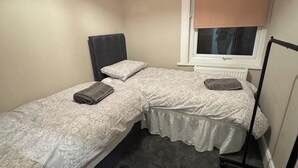 Un alojamiento pequeño y de una sola habitación con dos camas, una ventana con persiana, un radiador y un soporte de metal negro.