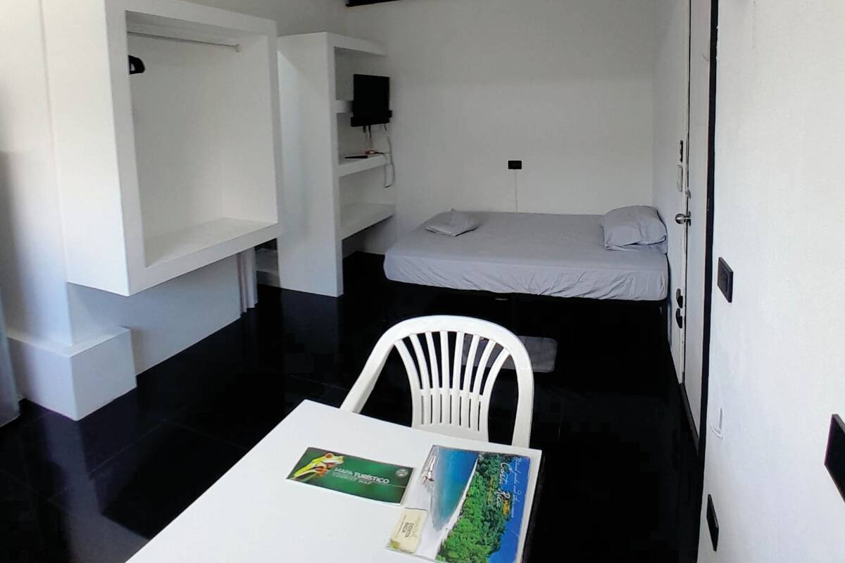 Quarto compacto com cama, uma cadeira branca, uma mesa pequena com um mapa e um ventilador de teto.