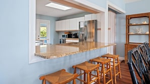 A kitchen with a central island, bar stools, and a view into the adjacent dining area. - Charming 4BR Cottage | Walk to Beach | Outdoor Space | Atlantic Beach (Atlantic Beach)