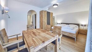 Un dormitorio con cama de madera, una mesita de noche, una mesa auxiliar, una mesa de comedor de madera con sillas y una lámpara fijada a la pared.