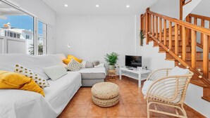 Una sala de estar moderna con un sofá, un reposapiés, sillones de mimbre, un televisor y una escalera de madera.