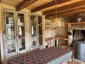 A rustic room with a wooden ceiling, a table with a patterned tablecloth, a hanging lamp, a fireplace, and a stone wall.