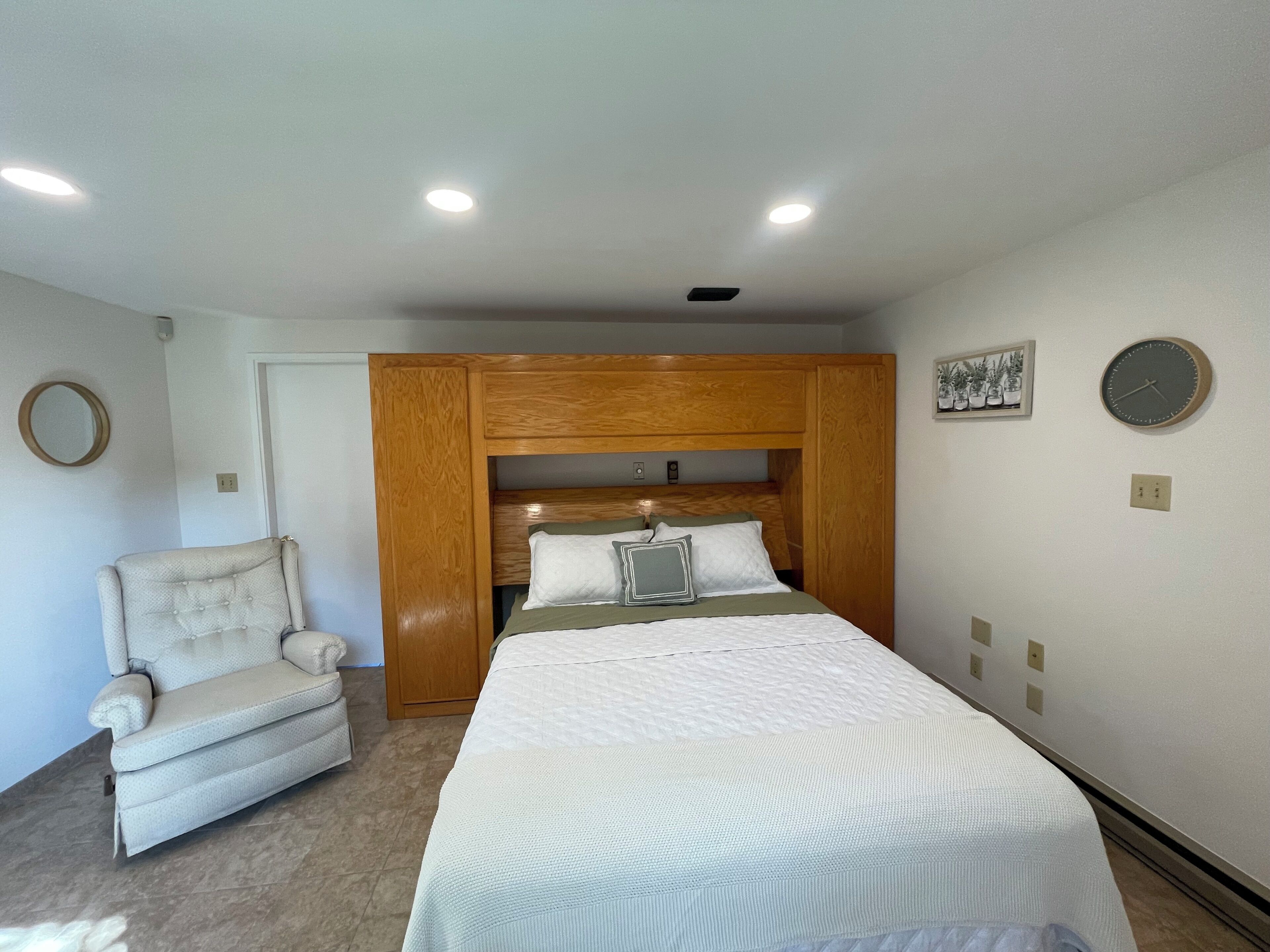 Un dormitorio con cama, una silla, cabecera de madera, un reloj de pared y un cuadro enmarco.