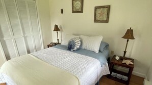 Une chambre à coucher comprenant un lit, deux tables de chevet, une commode, une fenêtre avec des stores et un mur orné de deux tableaux encadrés.