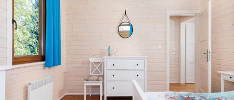 A room with wooden walls, a bed with a colorful bedspread, a white dresser, a chair, and a window with blue curtains.