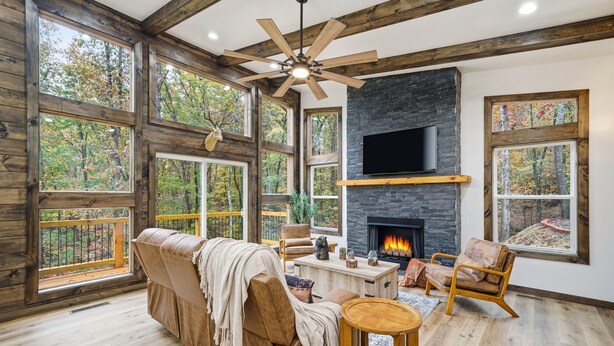 A spacious living room with a fireplace, wooden beams, large windows, and a ceiling fan. - Serene Ellijay Getaway with Coffee Bar, Two Fireplaces, Hot Tub, Games  + Resort (Ellijay)