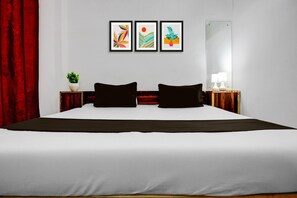 A hotel room with a large bed, two black pillows, a nightstand with a lamp, a mirror, and three framed artworks on the wall. - Hotel O Naroli Crossway Mathura (Mathura)