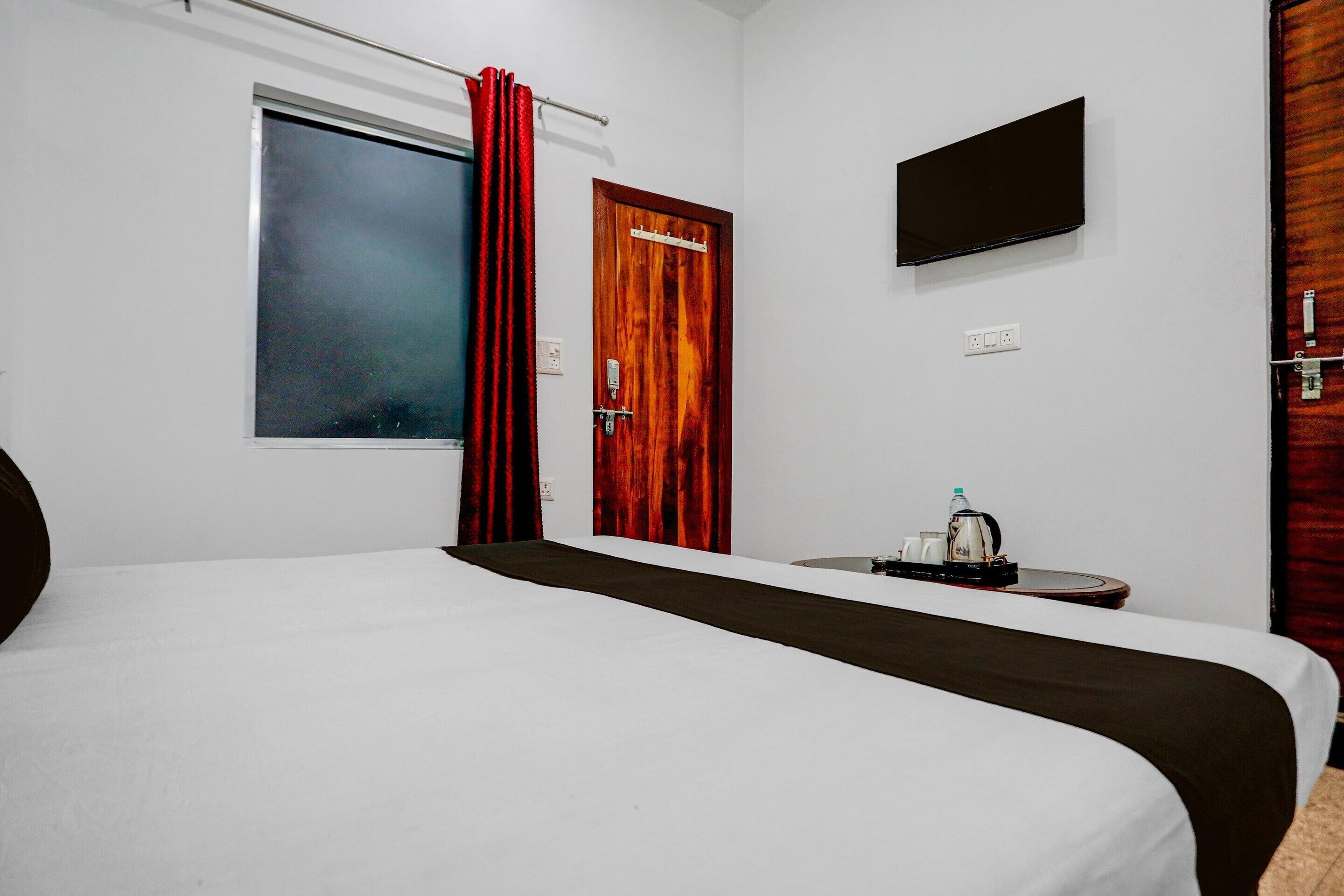 A hotel room with a bed, a TV mounted on the wall, a window with curtains, and a small table with a kettle and cups.