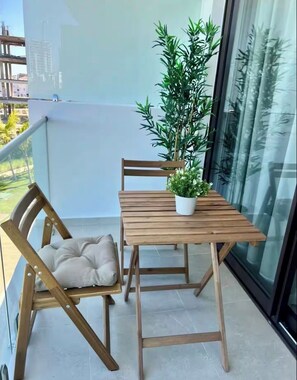 Een balkon met een houten tafel, twee stoelen, een bloempot en uitzicht op gebouwen.
