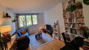 Un salon avec un canapé, une table basse et une bibliothèque. Il y a une fenêtre avec des rideaux, un radiateur et une lampe.