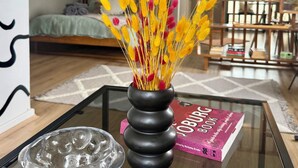 A living room with a glass coffee table, a black vase with yellow and pink decorative elements, a book titled 'BOURJOIS,' and a television in the background. - Jozi Muse: Retro Oasis in the Heart of Sandton (Sandton)