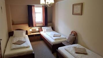 Habitación de hotel con dos camas individuales, una mesita de noche con lámpara, una silla, un ventanal con cortinas y un candelabro.