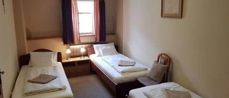 Habitación de hotel con dos camas individuales, una mesita de noche con lámpara, una silla, un ventanal con cortinas y un candelabro.