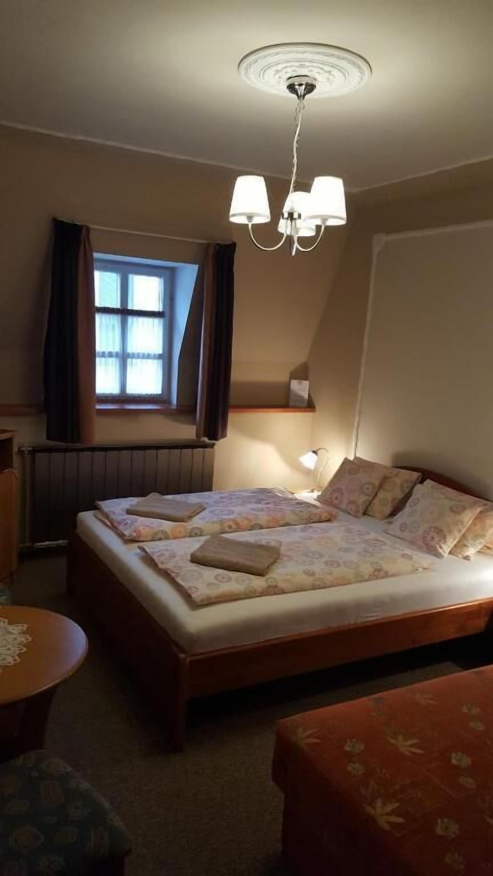 Un dormitorio con cama, una mesita, una silla, una ventana con cortinas e iluminación en el techo.
