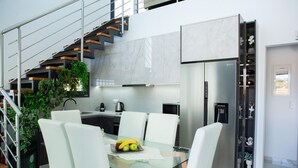 Une cuisine moderne avec une table à manger en verre, des chaises blanches, une cuisinière encastrée et un réfrigérateur en acier inoxydable.