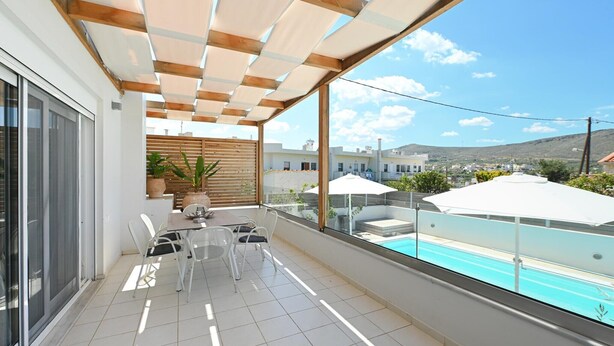 Een balkon met glazen hekwerk, een tafel met stoelen, een bloempot en uitzicht op een zwembad en gebouwen.