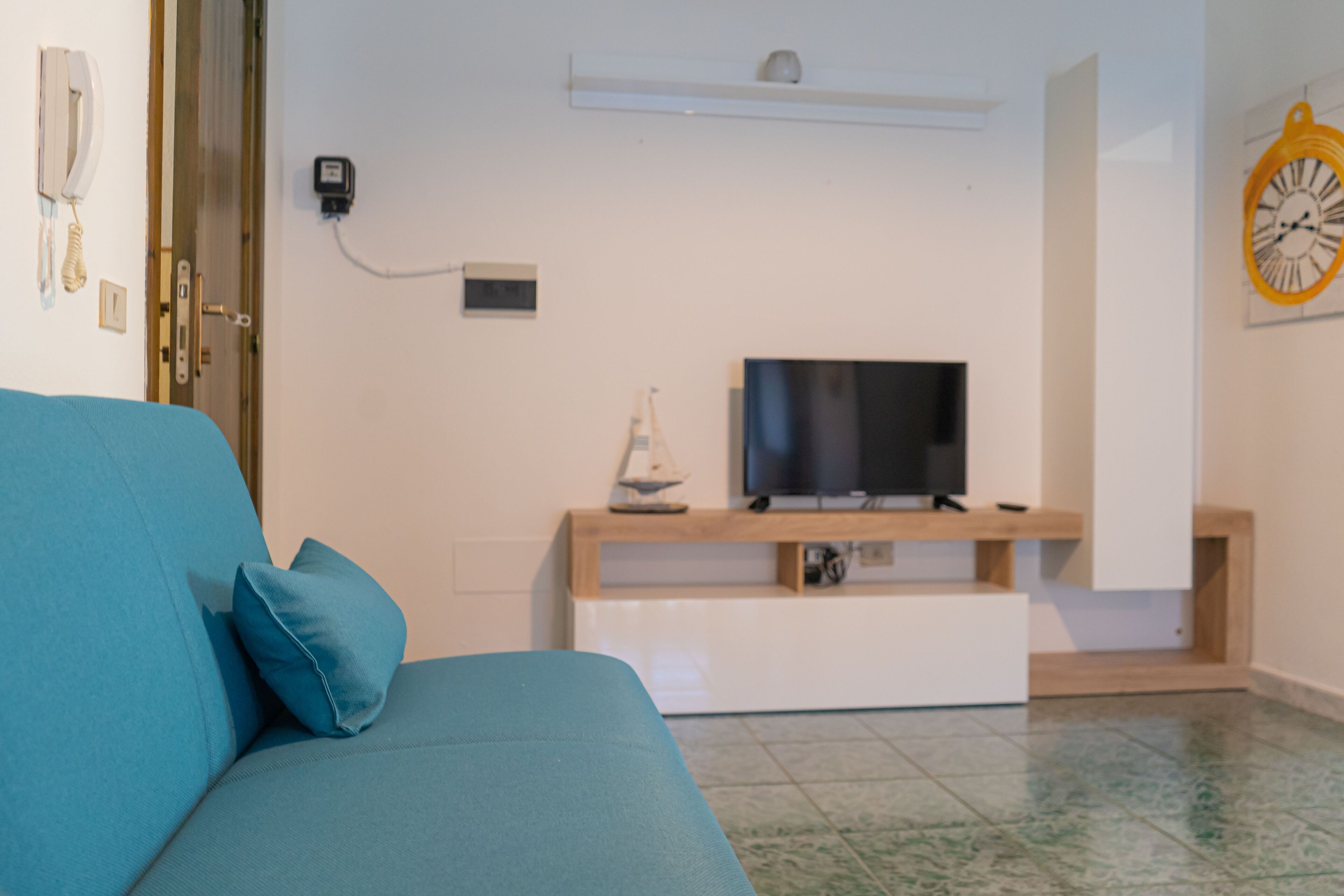 A living room with a sofa, a television on a stand, a wall clock, and a door with a phone.