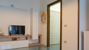 Quarto com televisão de tela plana em suporte de madeira, relógio na parede e porta que dá para outro espaço.