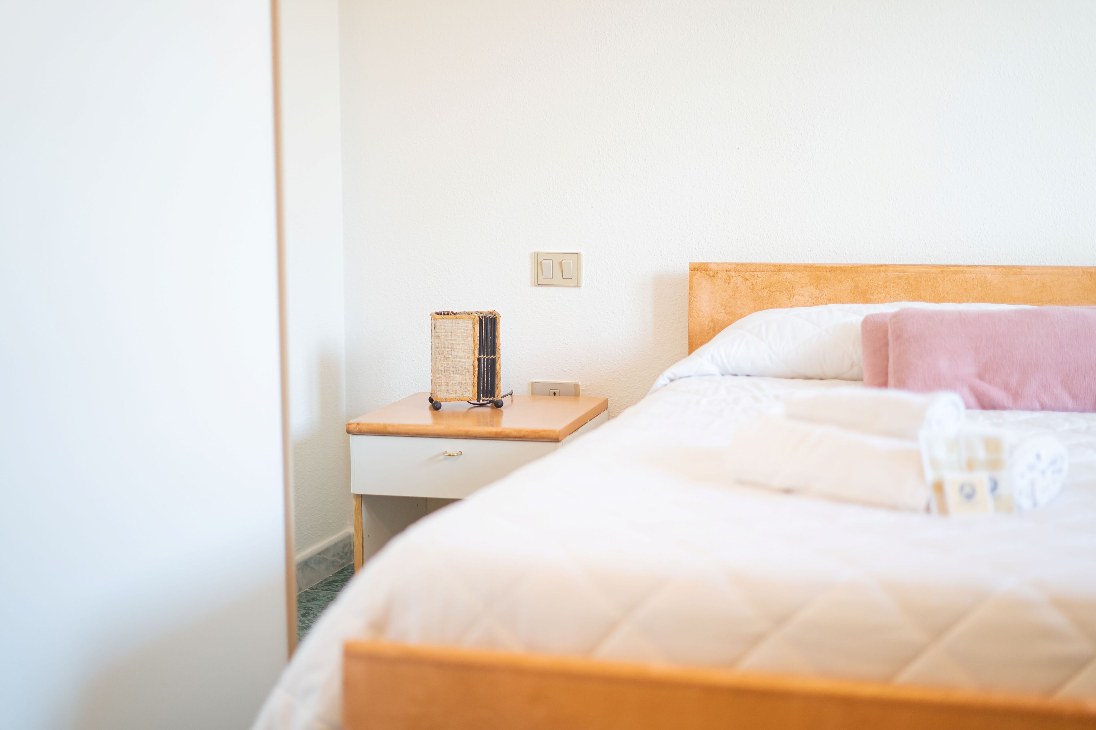 Una cama bien hecha con cabecera de madera, una mesita de noche con lámpara y un enchufe de pared.