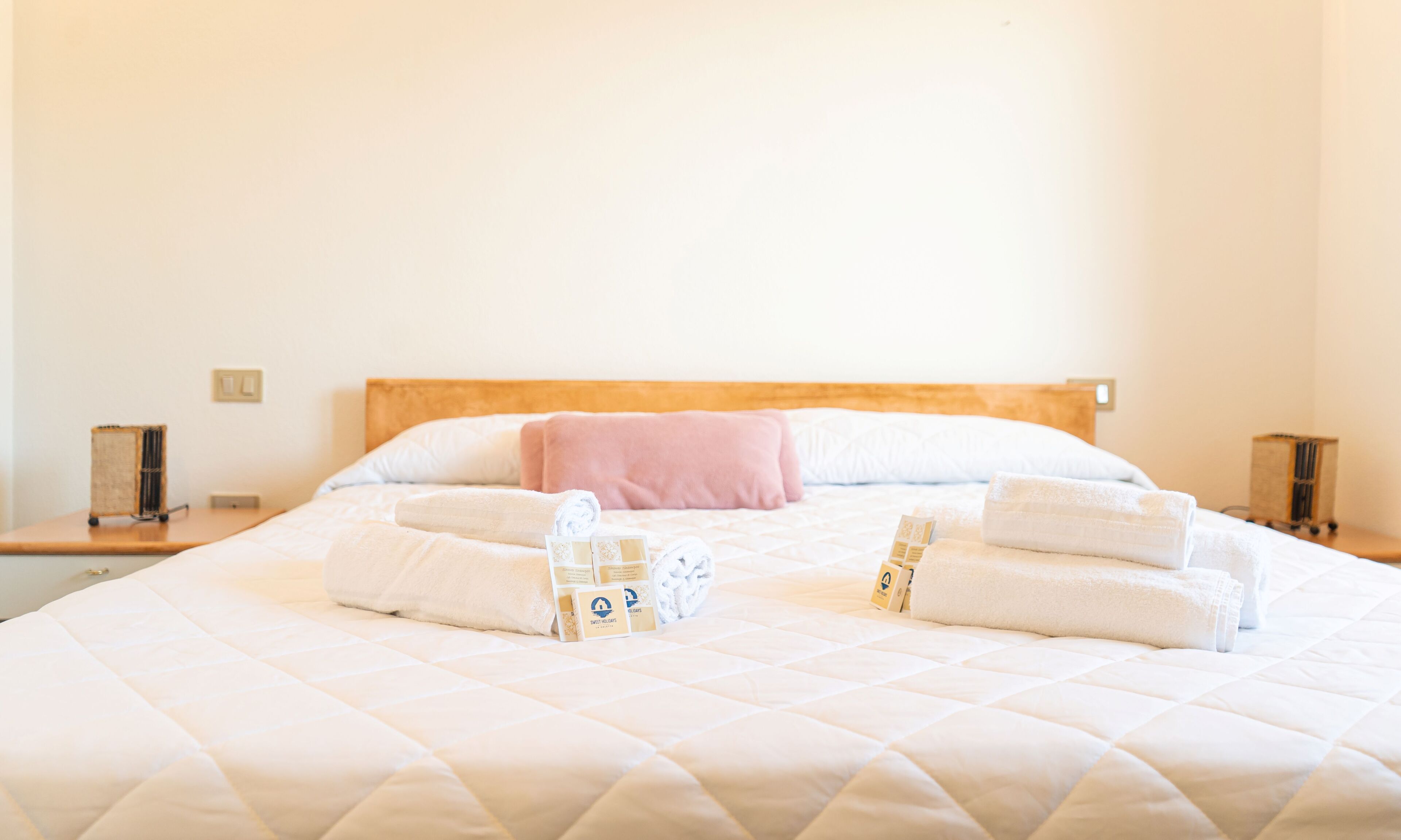 A neatly made bed with a quilted white duvet, a wooden headboard, and a pink pillow. There are two bedside tables with lamps and a small box of toiletries.