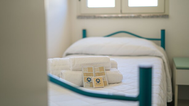 Uma cama com roupa de cama branca e cabeceira verde-água, uma janela com persianas e uma pequena mesa de cabeceira.