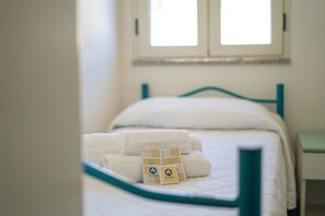 A bed with white linens and a teal headboard, a window with blinds, and a small bedside table. - Sweet Holidays Relax a La Caletta - Berchida 1 (La Caletta)