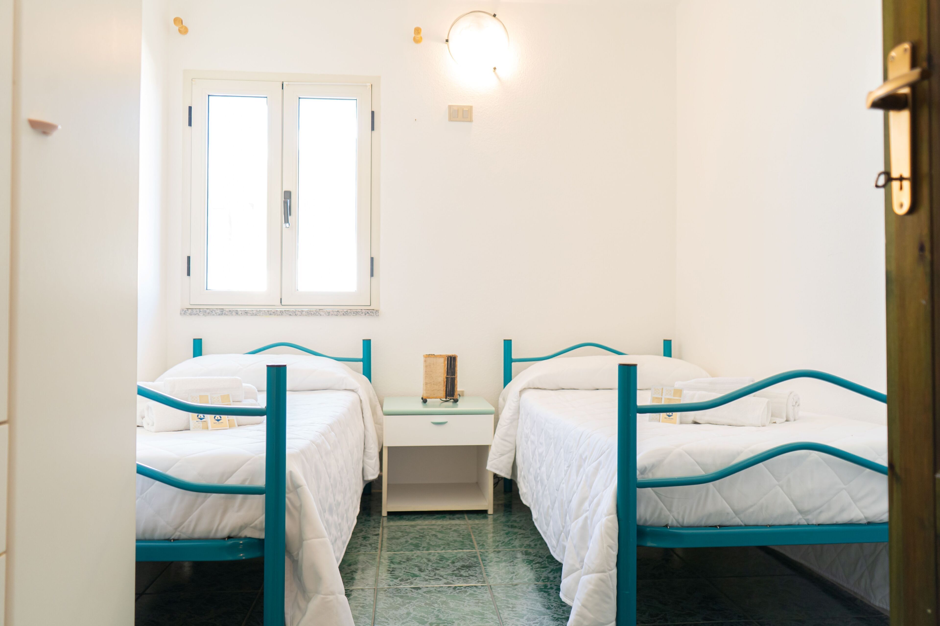 Une chambre avec deux lits, une table de chevet, une fenêtre et un luminaire fixé au mur.