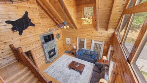 Interior de uma cabana de tronco com lareira de pedra, um sofá, uma mesa de centro e uma janela grande.