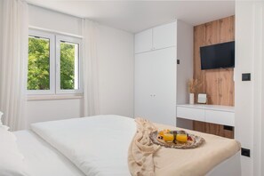 Ein Schlafzimmer mit Bett, einem Fenster mit Vorhängen, einem an einer Holzwand montierten Fernseher und einem Frühstücksbrettchen auf dem Bett.