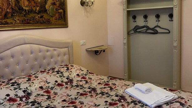 Een slaapkamer met een gestoffeerd hoofdeinde, een bloemenbedover, een kroonluchter, een aan de muur hangend schilderij, een kaptafel met spiegel en een kapstok.