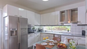 A modern kitchen with white cabinets, stainless steel appliances, and a dining area with a glass table set for breakfast. - 10-Guest Countryside Villa with Barbecue (Heraklion)