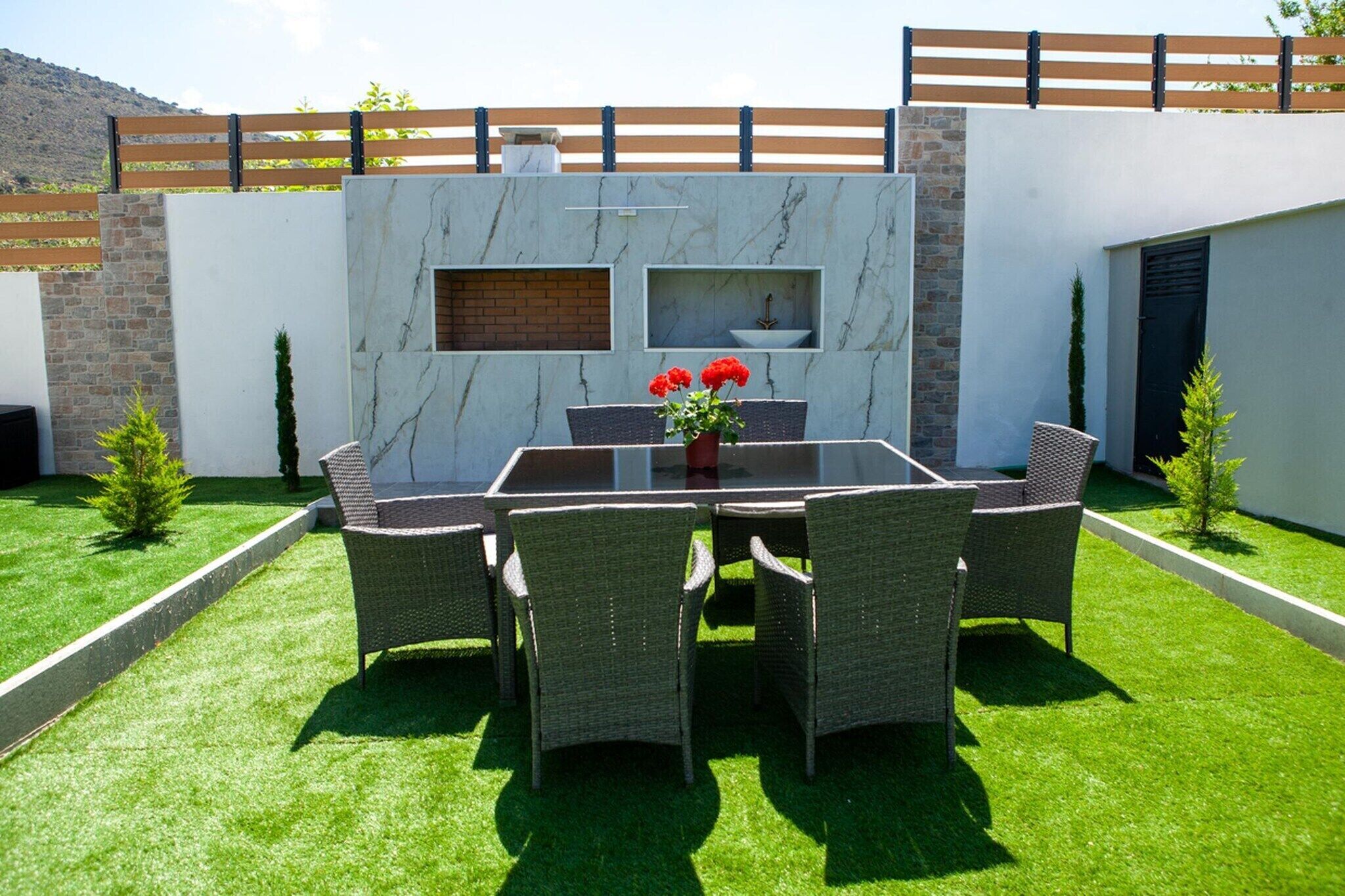 Eine Terrasse mit einem Glastisch, Korbstühlen und einem Blumentopf mit roten Blüten.