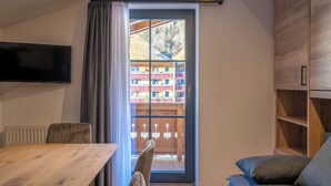 Una habitación con una mesa de comedor de madera, un sofá, un televisor montado en la pared y un ventanal corredero que da a un espacio exterior con vistas a edificios y una valla de madera.