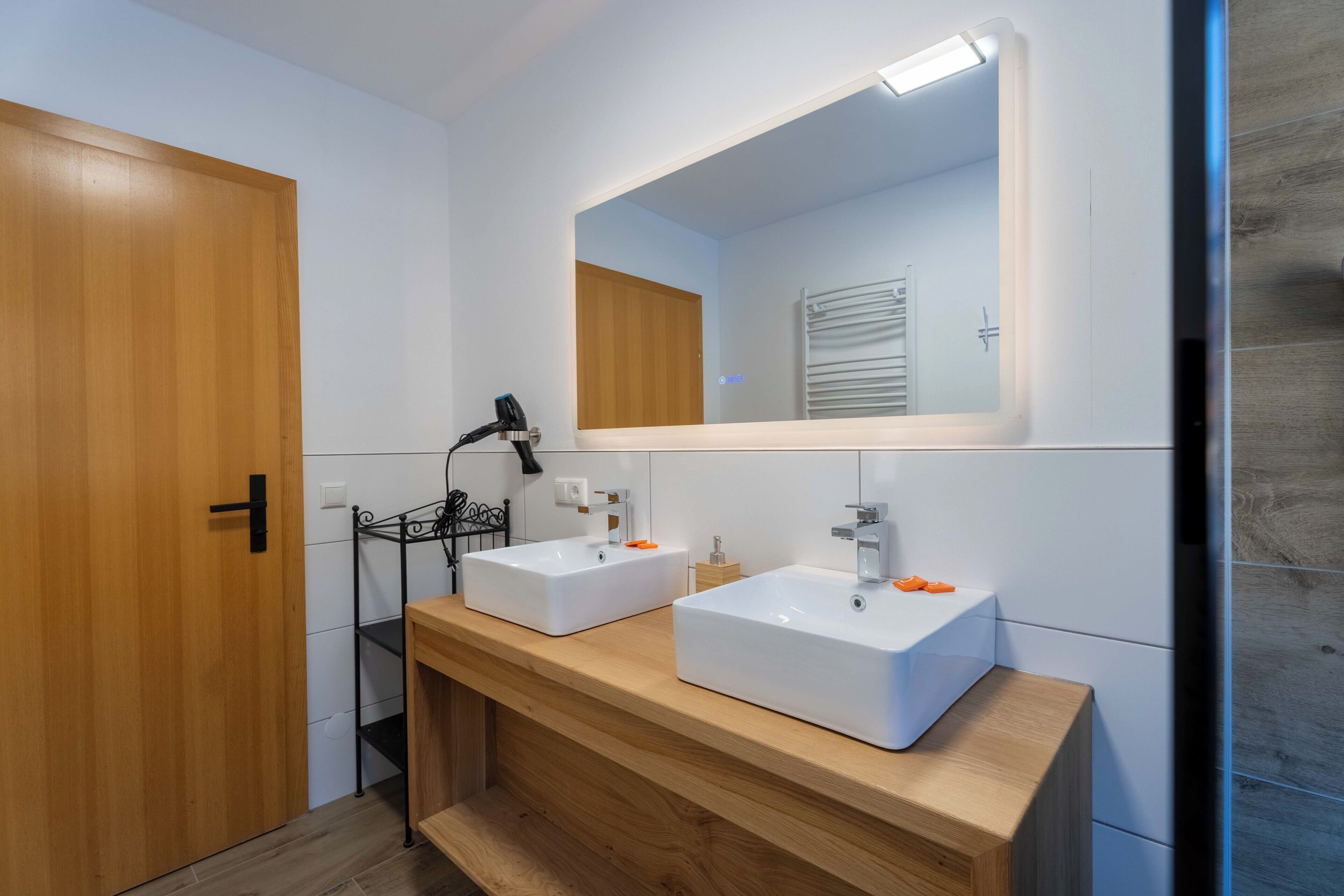 Un baño con dos lavamanos blancos, un mueble de tocador de madera, un espejo con iluminación integrada y una puerta.