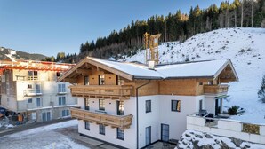 Una cabaña moderna con balcón de madera, un telesilla al fondo y un paisaje montañoso nevado.