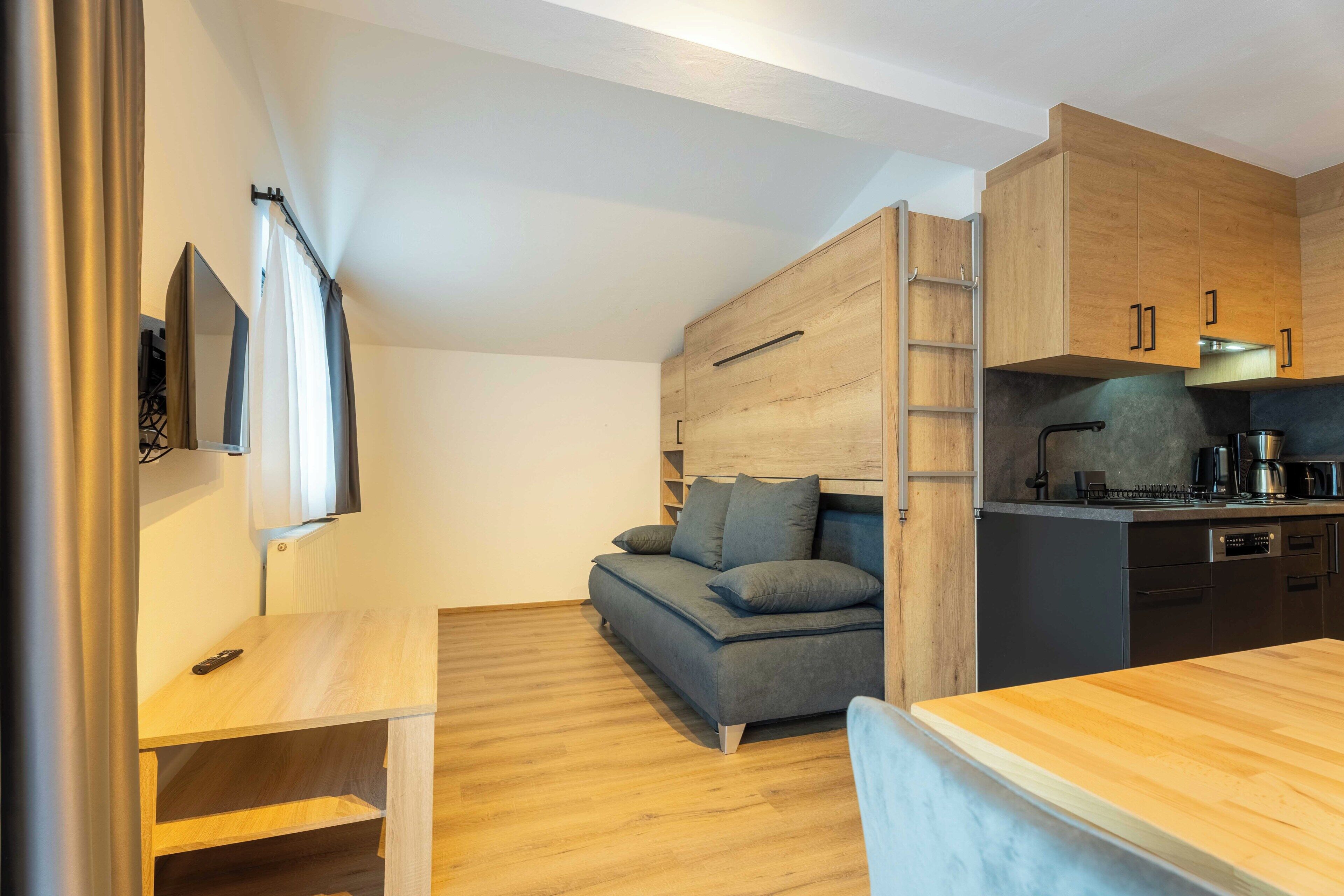 A compact living space with a sofa, a small table, a kitchenette, and a TV mounted on the wall.