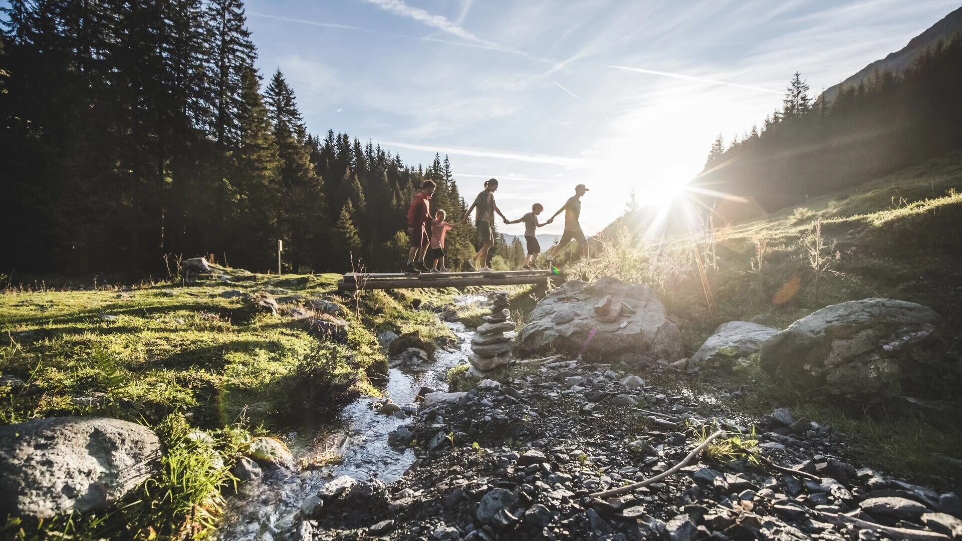 A group of people crossing a wooden bridge over a stream in a forested area with mountains in the background.
