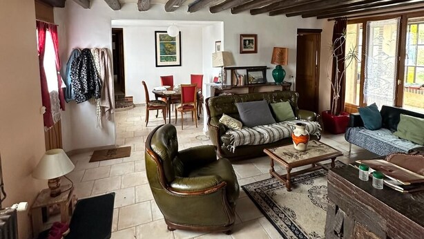 Un salon rustique avec des poutres apparentes, un fauteuil en cuir, un canapé avec des coussins, une table basse et, au fond, un coin repas.