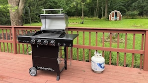 A Charbroil grill on a wooden deck with a propane tank nearby, surrounded by a wooded area. - 3-bedroom House, Private Lake Access and Game Room (Hillsboro)