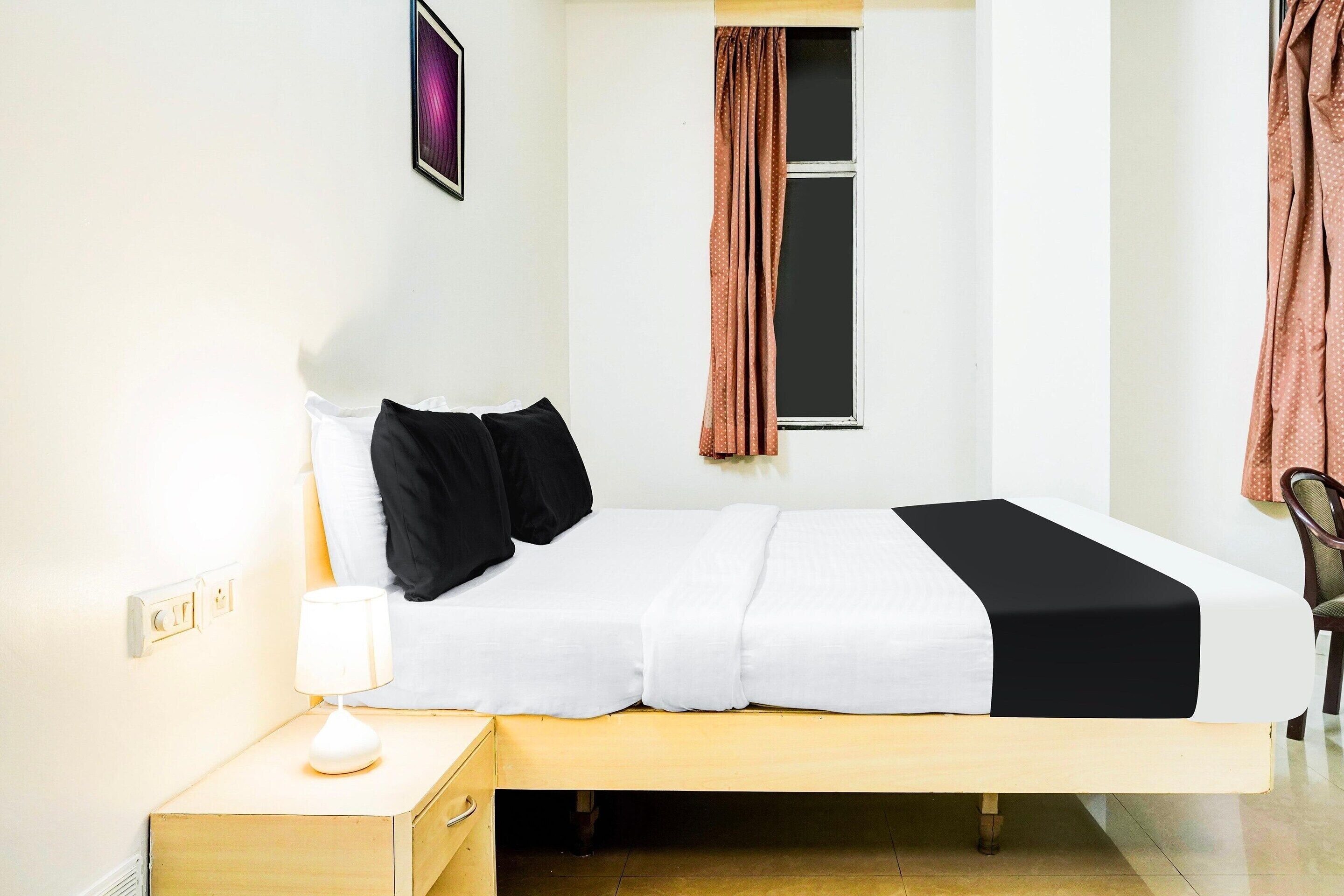 Una cama individual con colcha blanca y negra, una lámpara de noche, una ventana con cortinas y una silla.