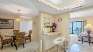 A modern living room with a dining area, a kitchen island, and a balcony with a view. - 🏖️Water Views | Steps to Beach | Beachfront Complex | Island Winds Penthouse 1 (Fort Myers Beach)