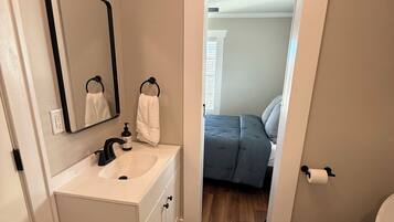 A bathroom with a sink, mirror, toilet, and a doorway leading to a bedroom.
