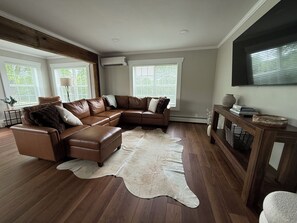 Un salon comprenant un canapé d’angle en cuir marron, un tapis en peau de vache blanche, un plancher en bois, une table d’appoint avec des livres et un vase, et un téléviseur fixé au mur.