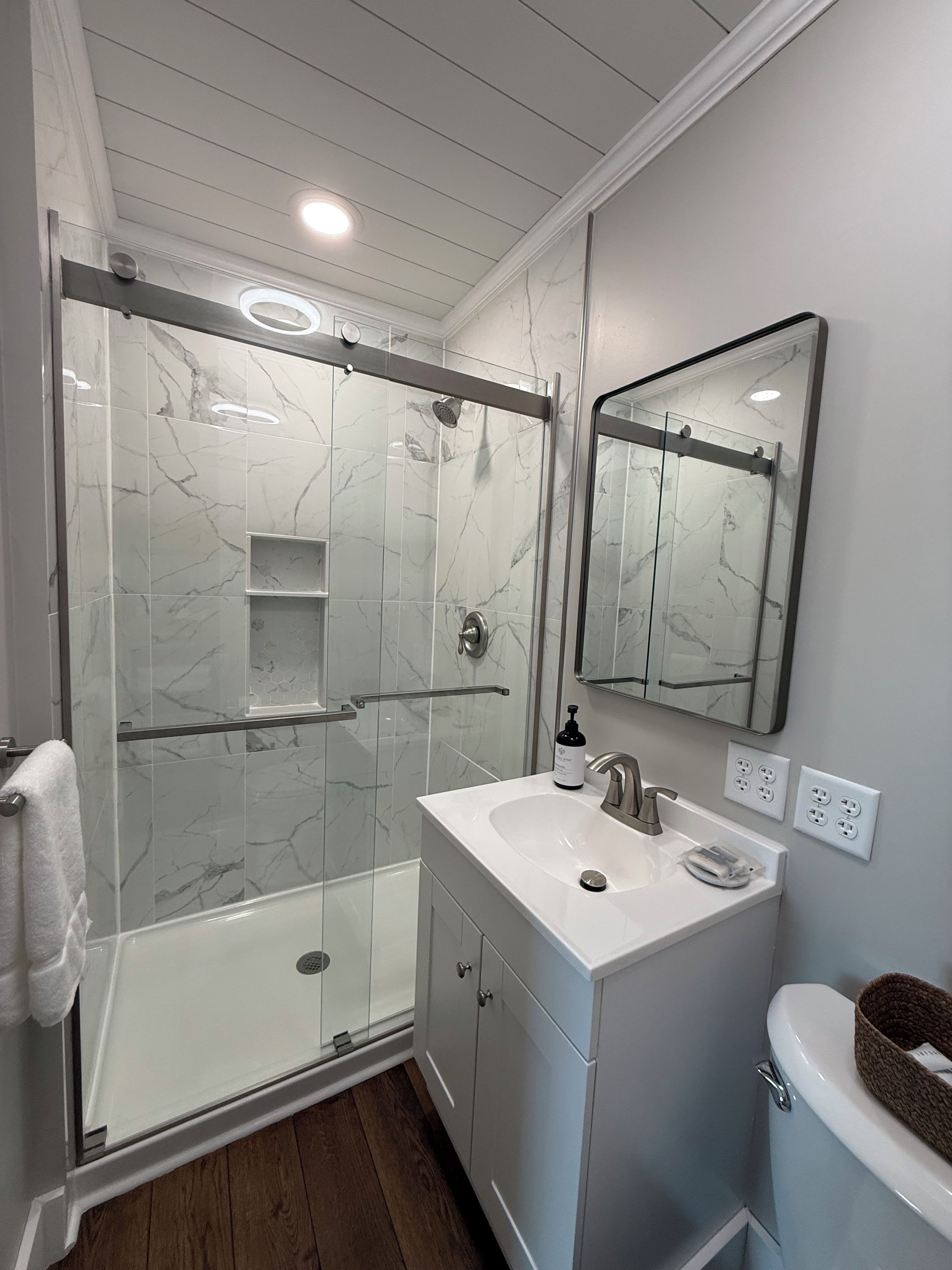 A modern bathroom with a glass-enclosed shower, a white sink with a mirror above, and wooden flooring.