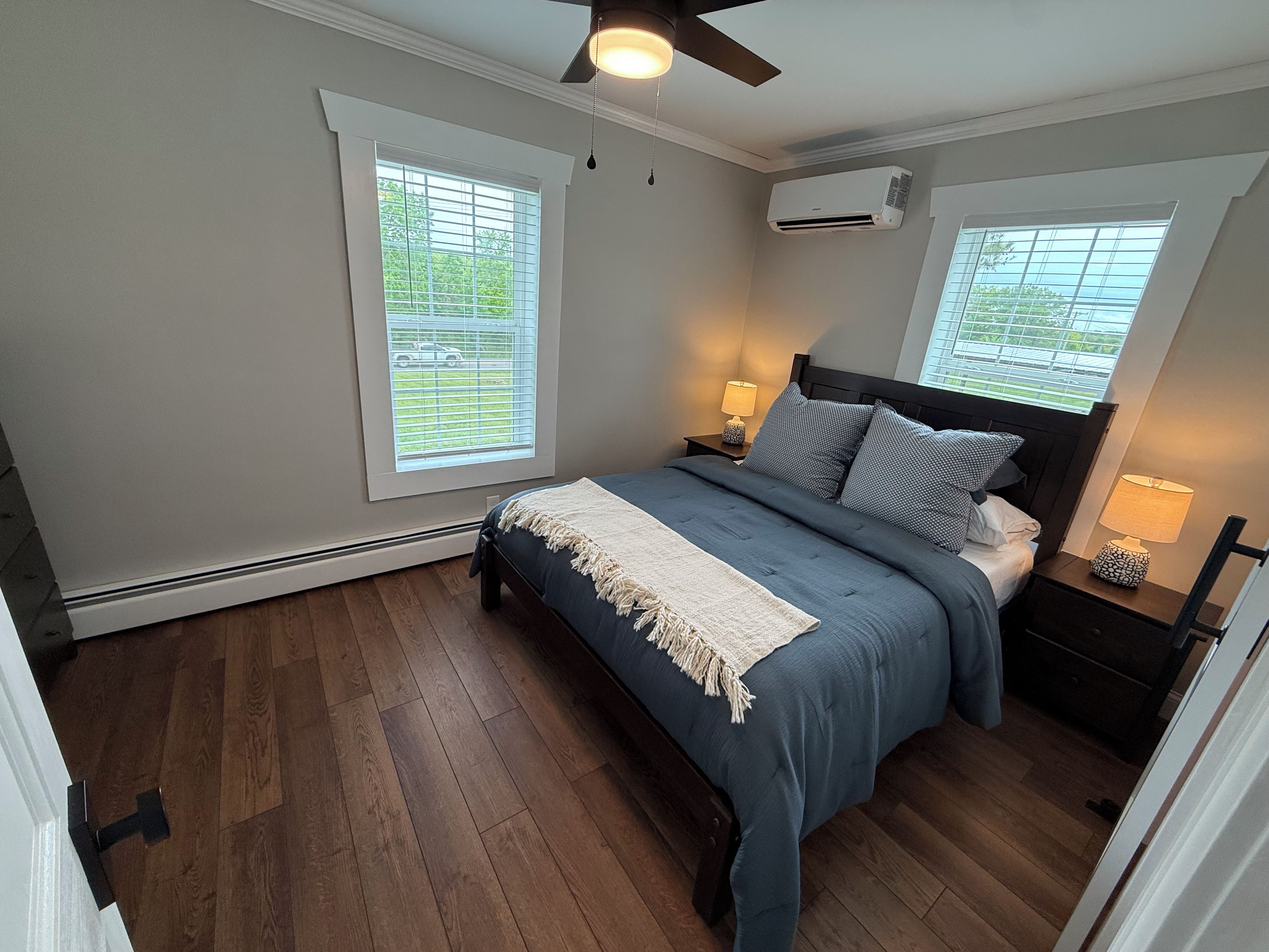 Un dormitorio con una cama, dos mesitas de noche, un ventilador de techo y dos ventanas con persianas.